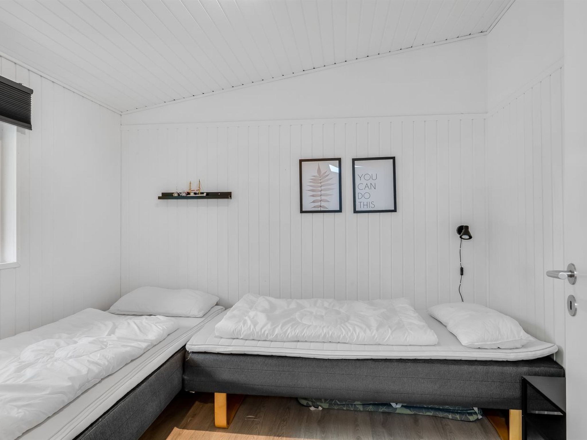 A room with two single beds, a small shelf with items, and two framed pictures on the wall.