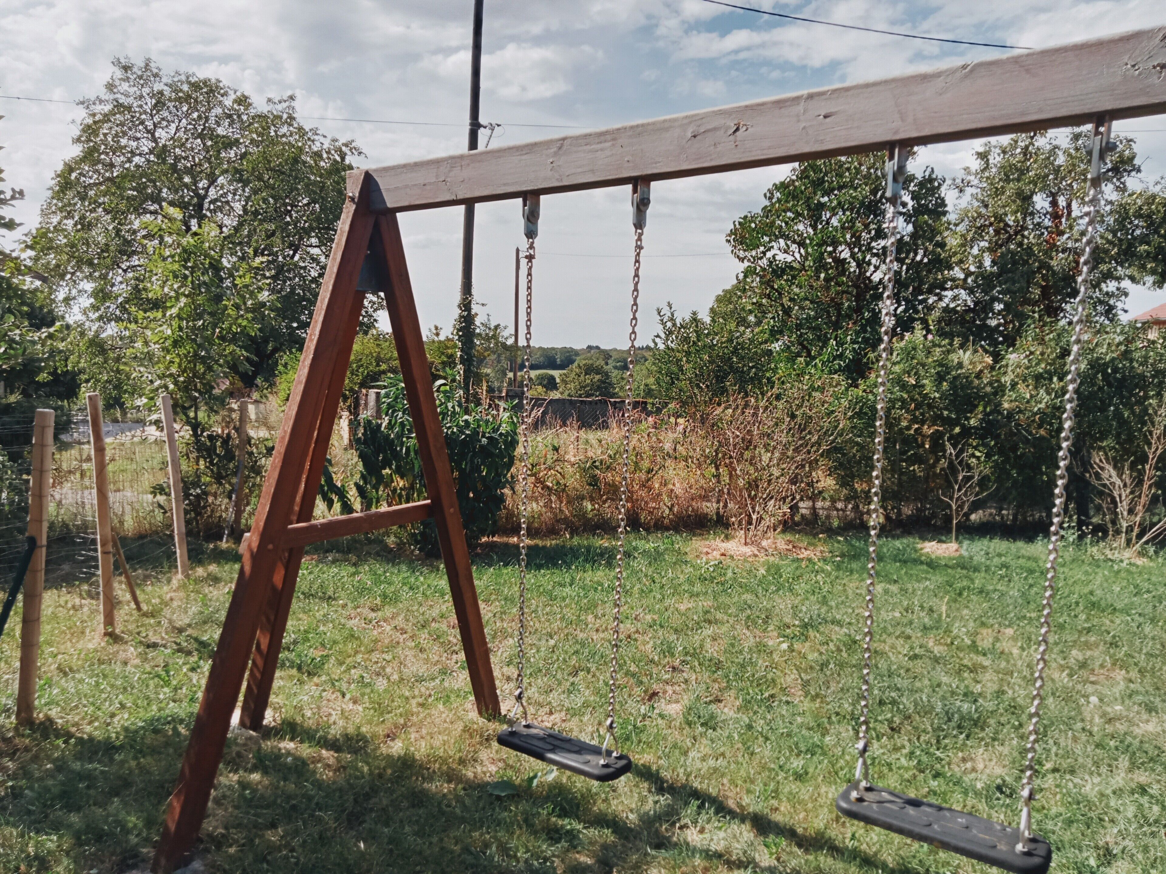 A wooden swing set in a grassy yard with trees and a fence in the background.