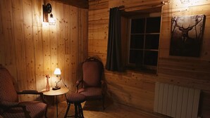A cozy cabin interior with wooden walls, a window with curtains, a radiator, and two patterned chairs around a small table with a lamp.