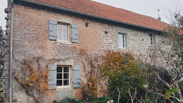 A stone building with two windows and shutters, surrounded by vegetation.