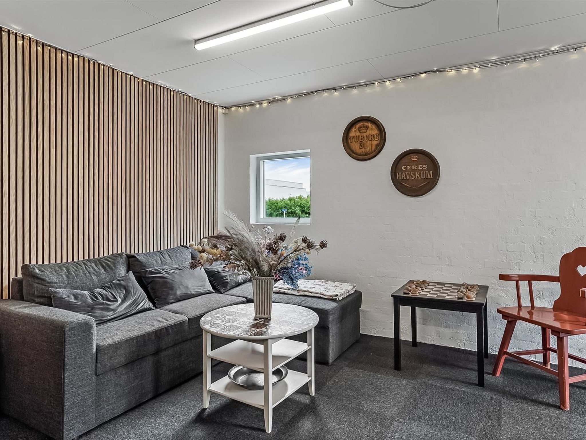 Una sala moderna con un sofá gris, una mesita redonda con un florero, una silla de madera y un tablero de ajedrez. Hay dos platos decorativos en la pared y una ventana con vistas a la vegetación.