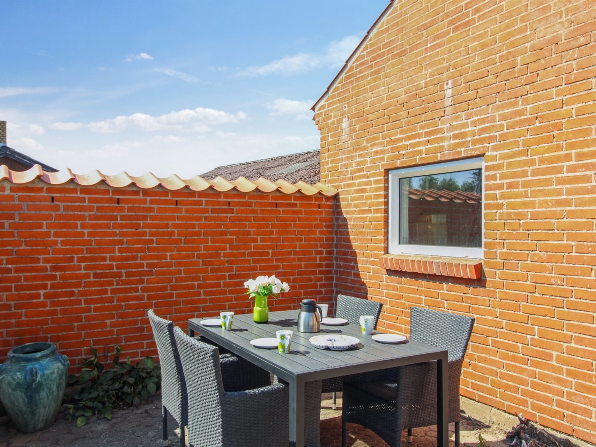 Un patio con una mesa preparada para cuatro personas, rodeado de sillas de mimbre, frente a una pared de ladrillo con una ventana.