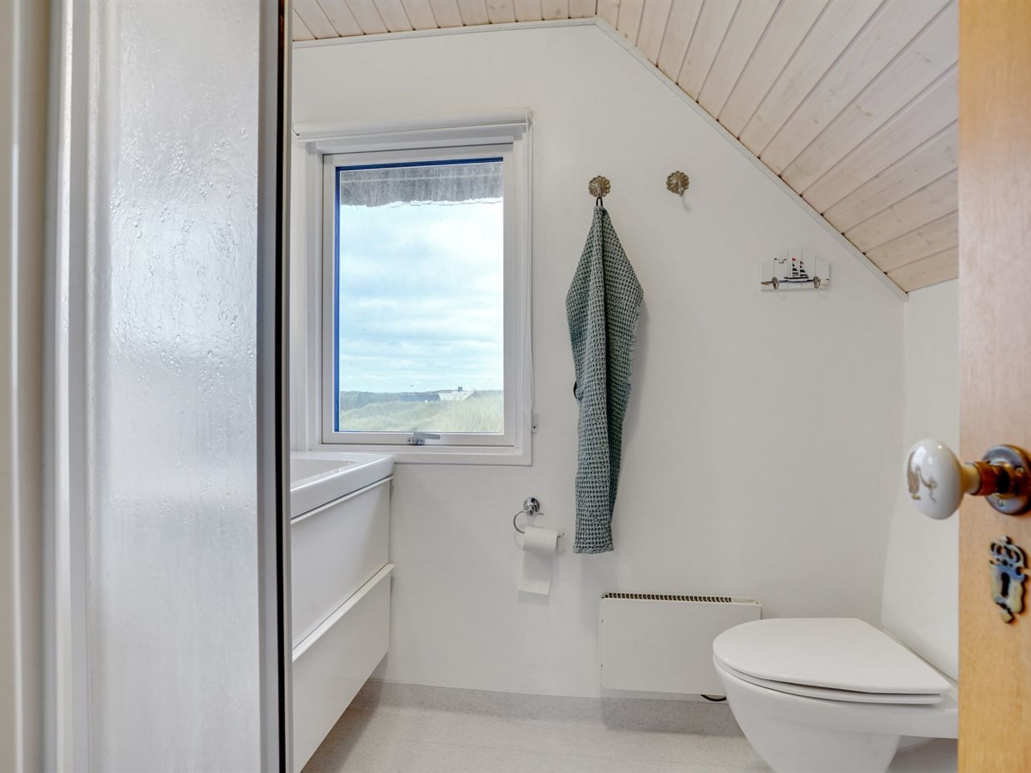 Et lille, hvidt badeværelse med et vindue, et håndklæde hængende på væggen og et toilet.