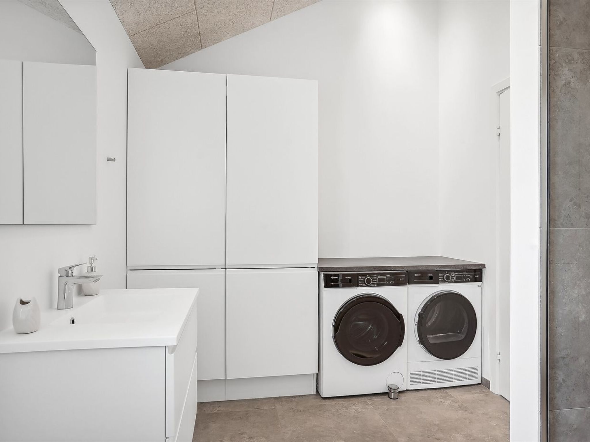 A laundry room with two front-loading washing machines, a white sink with a soap dispenser, and a large white cabinet.
