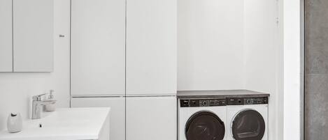 A laundry room with two front-loading washing machines, a white sink with a soap dispenser, and a large white cabinet.