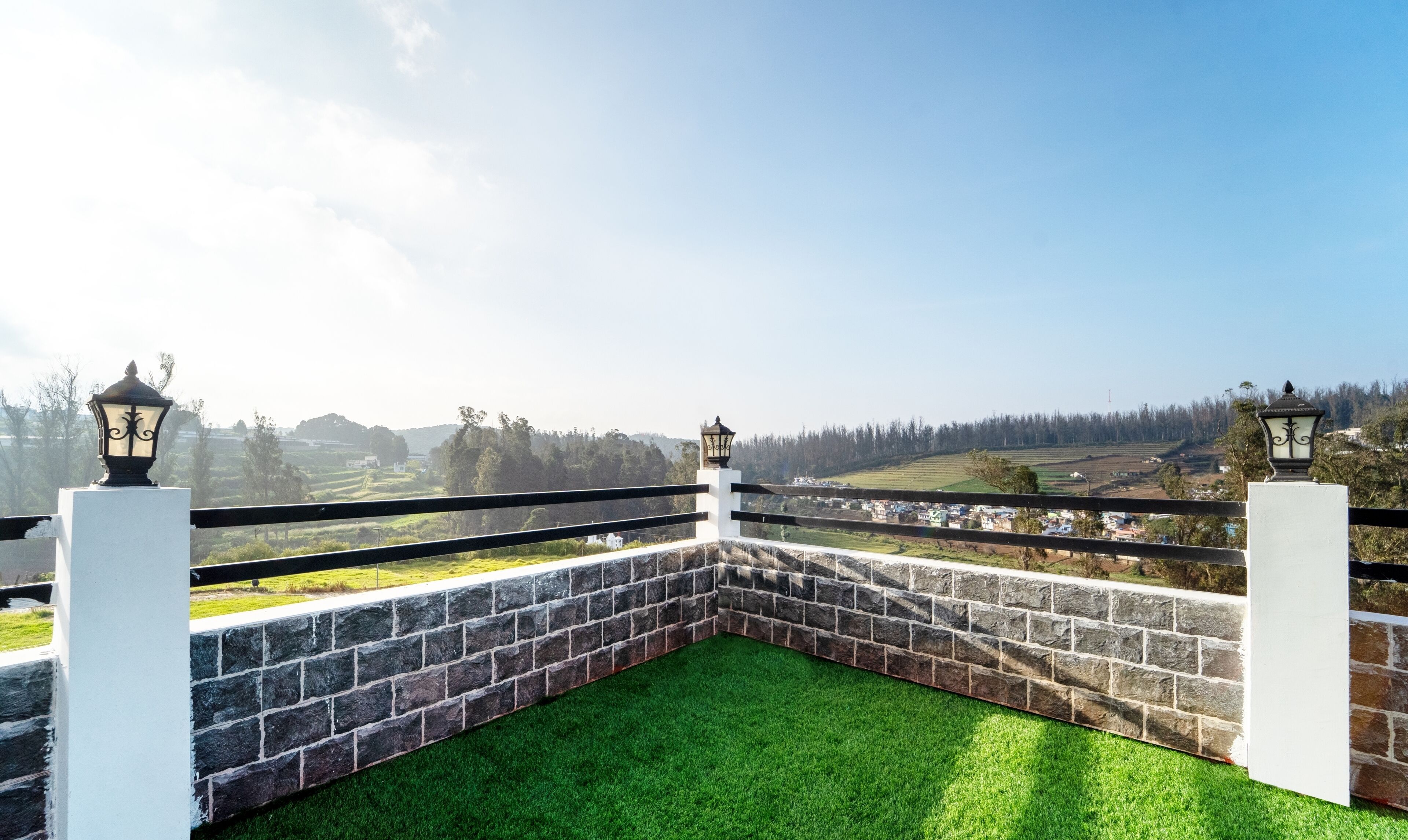 A balcony with a stone wall and green artificial grass, overlooking a rural landscape with fields and trees.
