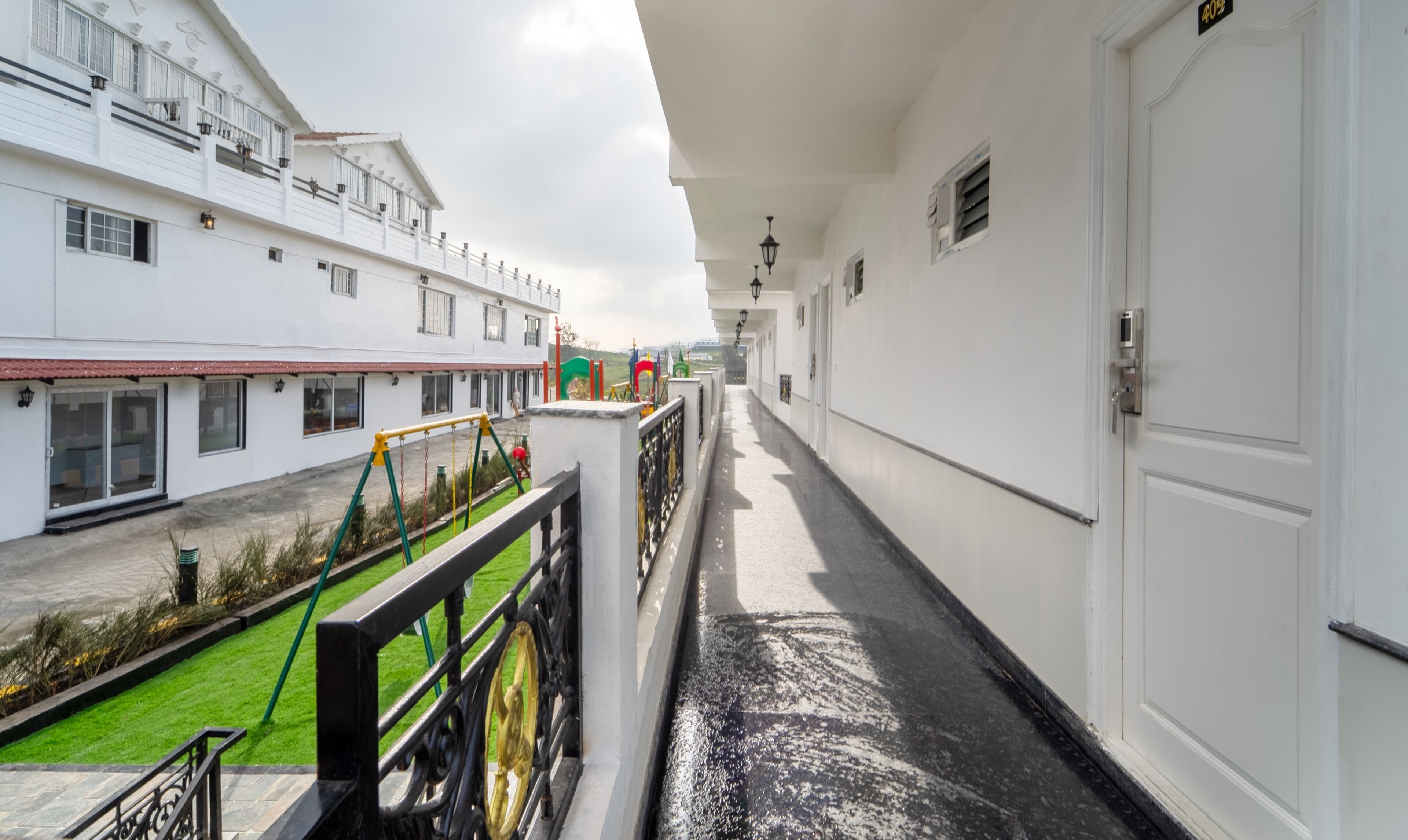 A corridor with a white building on one side and a fenced outdoor area with playground equipment on the other.