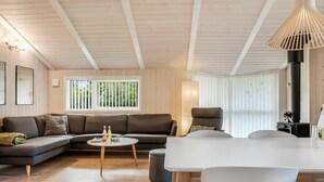 A modern living room with a sectional sofa, a round coffee table, and a dining table with chairs. There is a large window with blinds, a fireplace, and a ceiling with exposed beams. - "Barnet" - 2.2km from the sea by Interhome (Blokhus)
