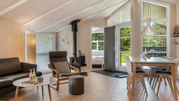A cozy living room with a fireplace, a sofa, a dining table with chairs, and a view of the outdoors through large windows. - "Barnet" - 2.2km from the sea by Interhome (Blokhus)