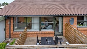 Een twee verdiepingen tellend huis met een golfplaten dak, grote ramen en een patio met een tafel en stoelen.