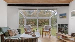Una sala de estar moderna con un gran ventanal que da a un jardín, un sofá, dos sillones, un televisor y suelo de madera.