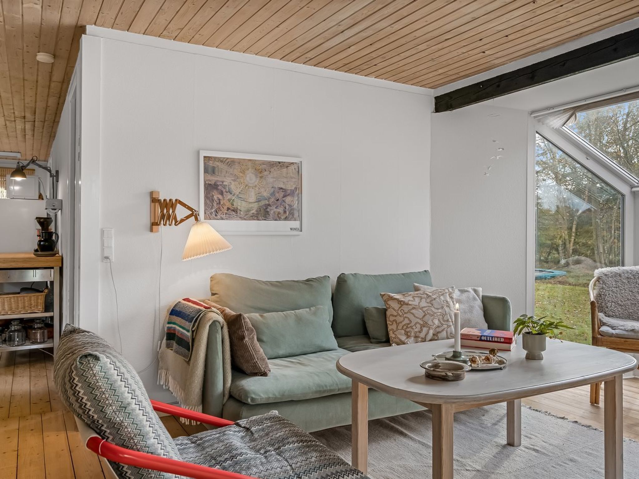 A modern living room with a sofa, a coffee table, and a chair. There is a painting on the wall, a lamp, and a window with a view of the outdoors.