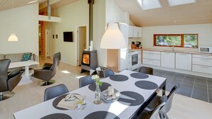 Une cuisine moderne avec un îlot central, un coin repas avec une table et des chaises, et un espace de vie avec un canapé et des fauteuils. Le plafond est doté de lucarnes et la cuisine est éclairée par une grande fenêtre.