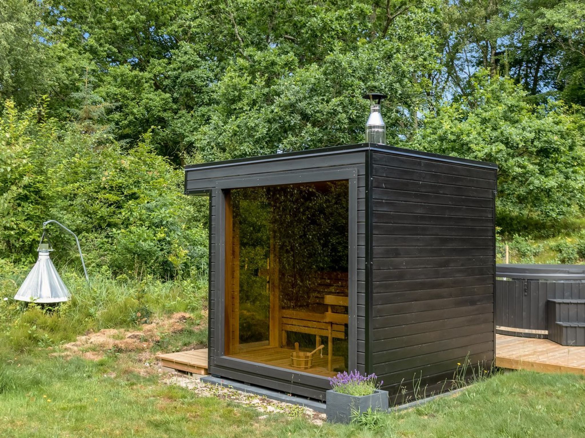 Una petita cabana de fusta fosca amb una porta de vidre, una banca de fusta a l'interior i una petita zona de jardí amb flors.