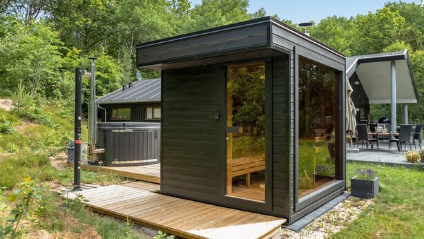 Une cabane moderne aux couleurs sombres, dotée d’une porte en verre, d’une terrasse en bois et d’un espace patio aménagé avec du mobilier d’extérieur.