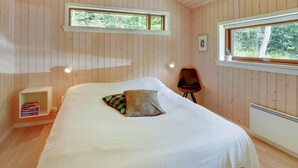 Une chambre avec des murs en bois, un lit avec une literie blanche, deux fenêtres, une chaise et une table de chevet avec des livres.