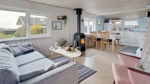 Een gezellige woonkamer met een bank, een eethoek met een tafel en stoelen, een keuken met witte kastjes, en een houtkachel.