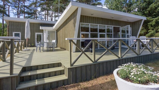 En moderne hytte med terrasse, udendørs møbler og et stort vindue med udsigt over den omkringliggende skov.