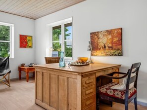Una habitación con una mesa tipo isla de madera, un cuadro de ciervos en un bosque, un tablero de ajedrez y una silla con cojín estampado.