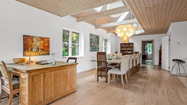 Una habitación amplia con techo de madera, un comedor con una mesa larga y sillas, y un gran ventanal que proporciona luz natural.