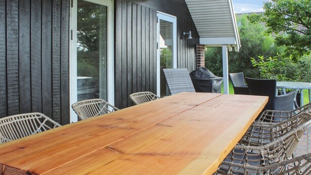 Een houten eettafel met rieten stoelen, buiten op een terras met uitzicht op een huis en groen.