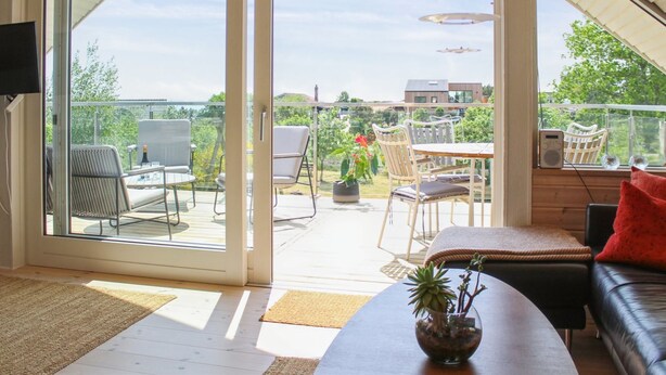 A spacious living area with a sliding glass door leading to an outdoor patio. Inside, there is a sofa, a coffee table with a plant, and a TV mounted on the wall.