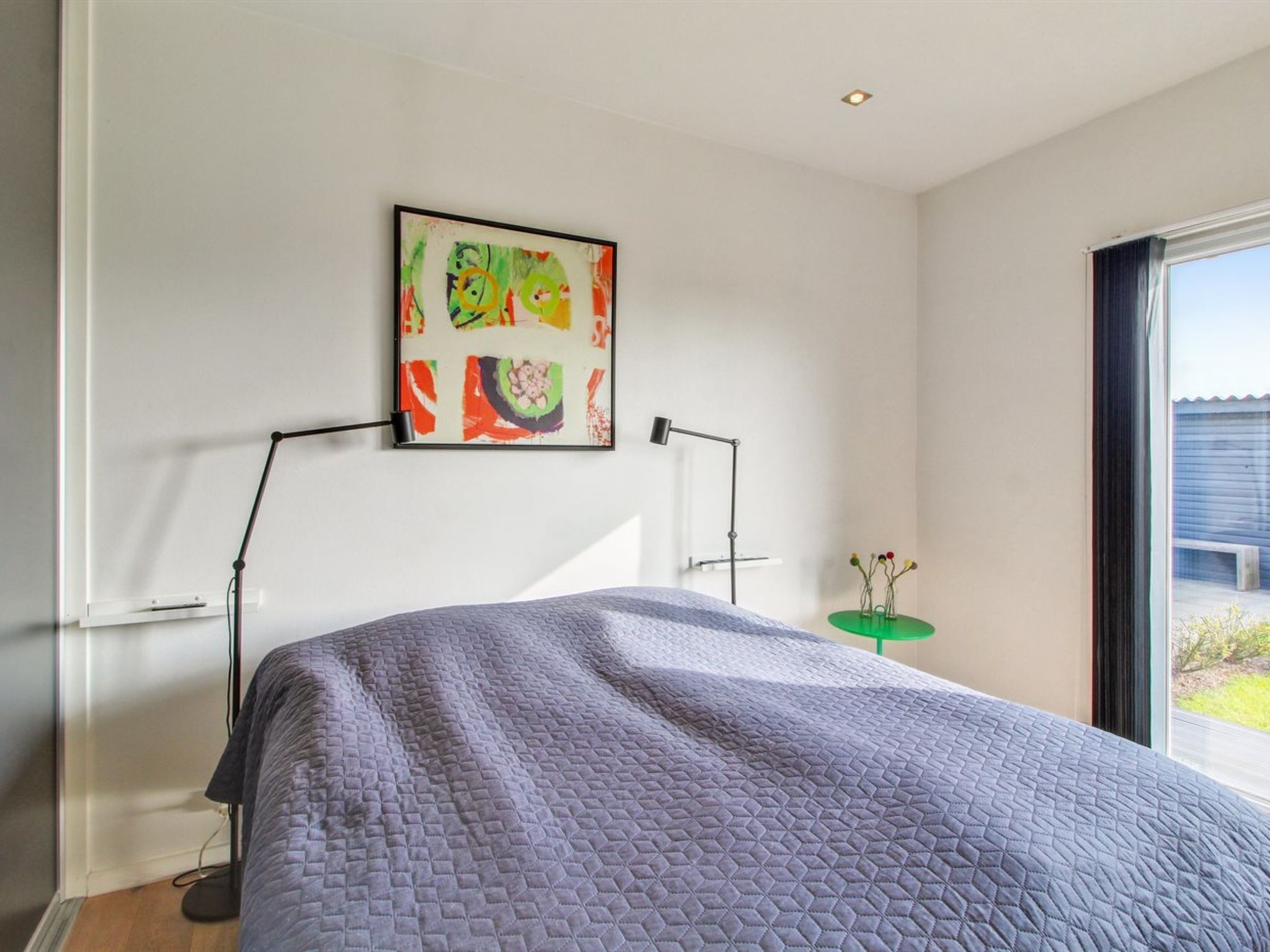 Une chambre moderne avec un grand lit, un tableau abstrait aux couleurs vives, une lampe sur pied et une vue dégagée sur l’extérieur grâce à une grande fenêtre.