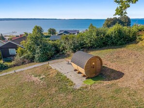 Een houten sauna op een grashelling, met uitzicht op water en huizen op de achtergrond.