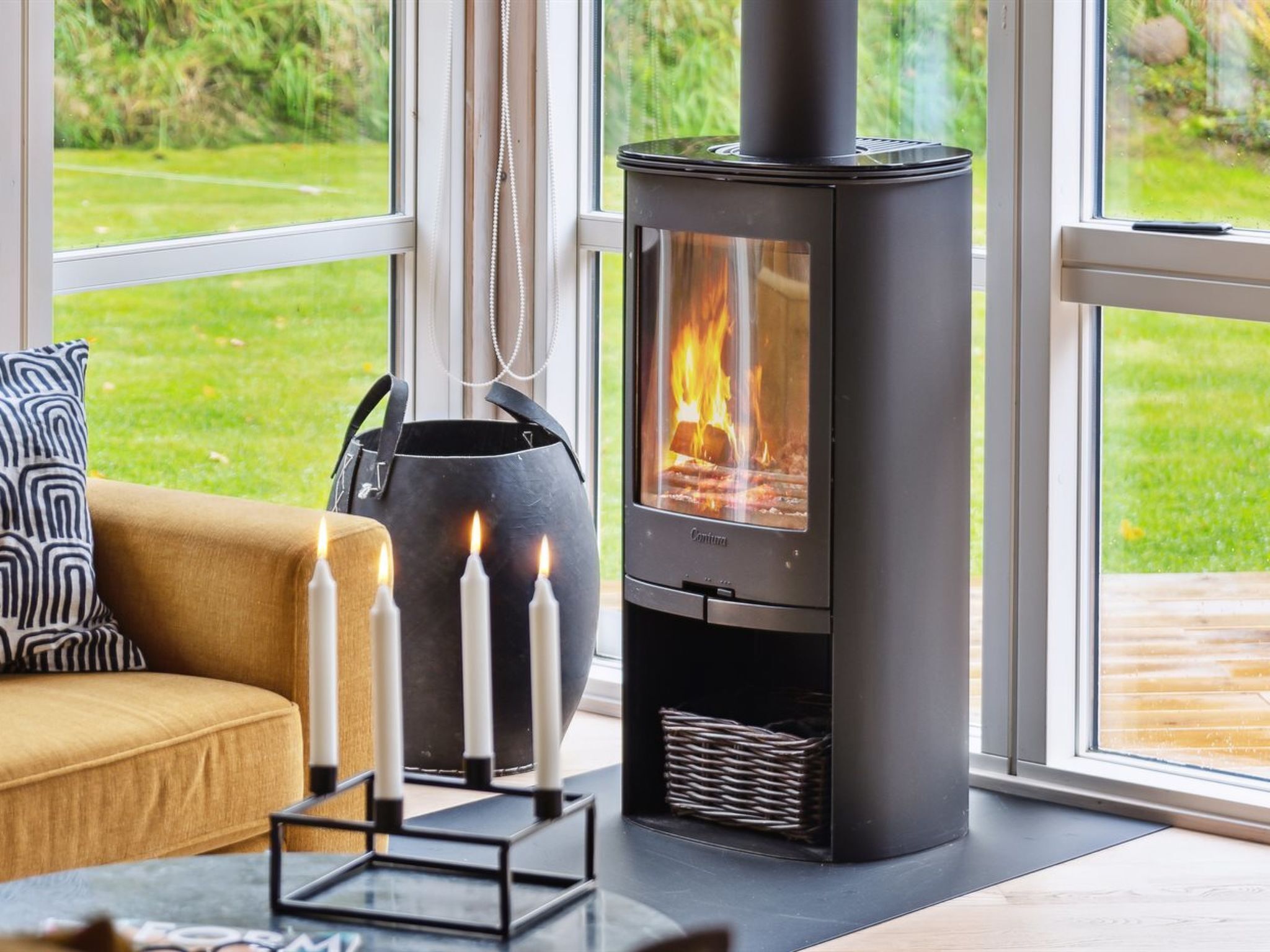 Un salon moderne avec un poêle à bois noir, un canapé beige et une grande fenêtre donnant sur un jardin verdoyant.