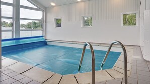 An indoor swimming pool with clear blue water, metal pool ladders, and a view of greenery outside.