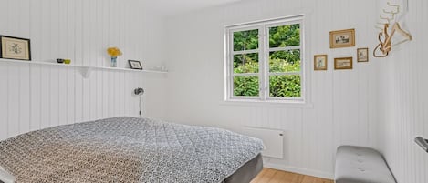 A bedroom with a bed, a window, a radiator, a shelf with decorative items, and a bench.