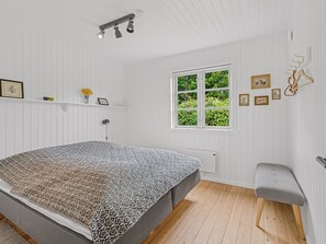 Quarto com cama, janela, radiador, prateleira com objetos decorativos e um banco.