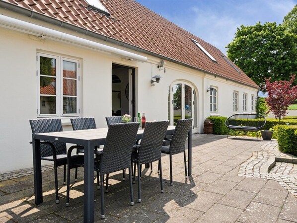Un bâtiment blanc avec un toit en tuiles, une terrasse avec une table et des chaises, et un jardin agrémenté d’un banc.