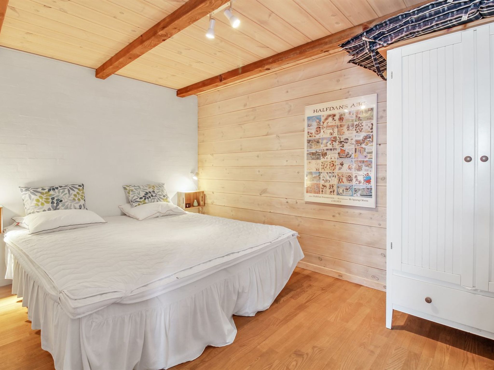 Un dormitorio con techo de madera, una cama grande con ropa blanca, un cartel en la pared y un armario blanco.
