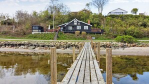 A wooden dock leading to a house with a dark exterior, surrounded by greenery and a stone wall.