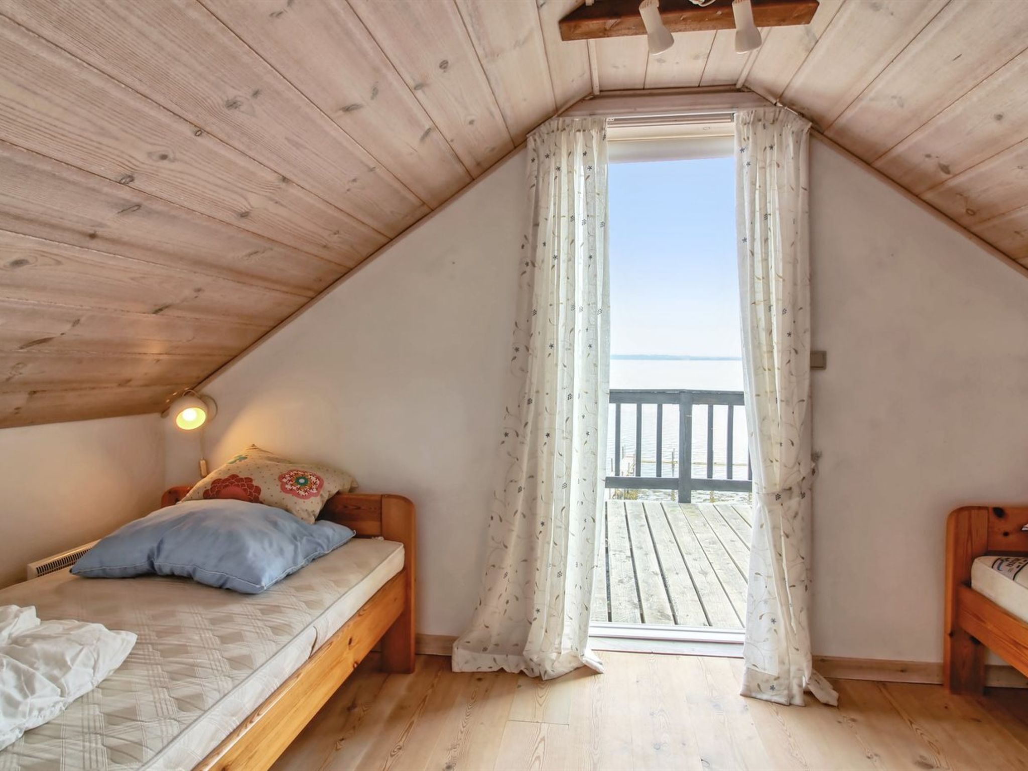 A small, cozy room with a sloped ceiling, a bed with a mattress and pillows, a bedside table, and a window with curtains.
