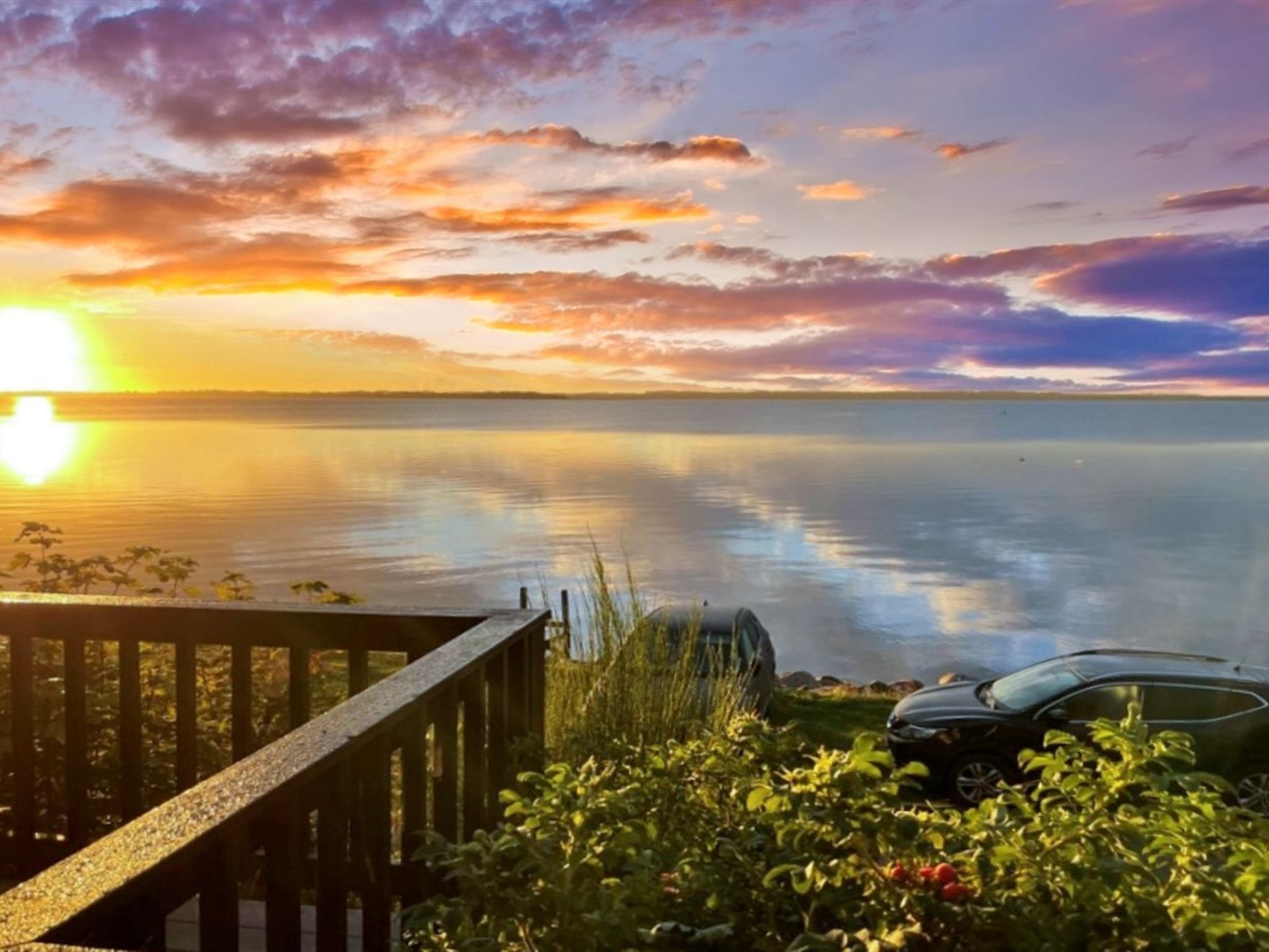 A serene lakeside view with a wooden deck and a parked car.