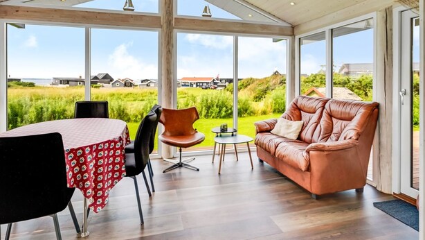 Une pièce spacieuse avec une grande table, un canapé en cuir et une vue sur un paysage verdoyant, visible à travers de grandes fenêtres.
