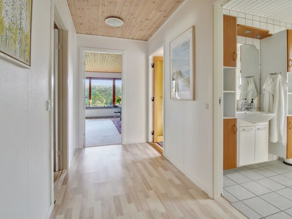 A hallway leading to a bathroom with a sink and mirror, wooden flooring, and a view of greenery outside.