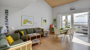 A spacious living area with a high ceiling, wooden beams, a dining area with a table and chairs, and a view of the sea through large windows.