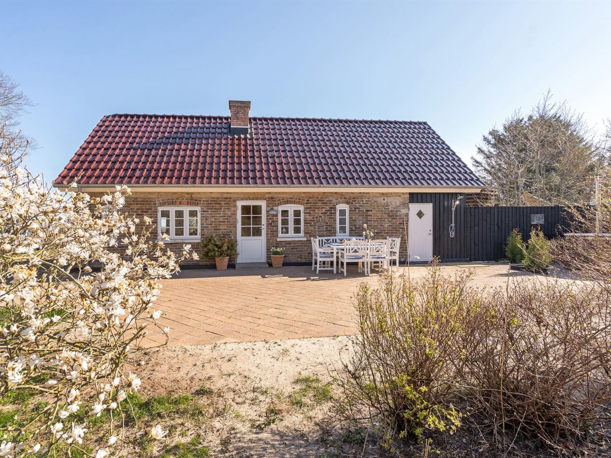 Une maison à deux étages, avec un toit recouvert de tuiles rouges, une façade en briques et une terrasse dallée aménagée avec du mobilier d’extérieur.