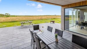 Een terras buiten met een tafel, stoelen, een houten bankje en uitzicht op een grasveld.