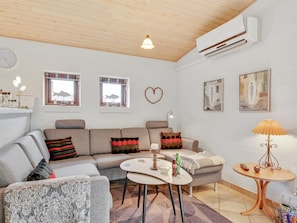 Ein modernes Wohnzimmer mit einem Ecksofa, einem runden Couchtisch und einem kleinen Beistelltisch. Zwei Fenster mit Vorhängen, eine Wanduhr und einige gerahmte Kunstwerke sind ebenfalls zu sehen.