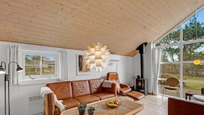 Un salon chaleureux avec un canapé en cuir, une table basse en bois sur laquelle sont disposés des fruits, un luminaire suspendu moderne et une grande fenêtre offrant une vue sur l’extérieur.