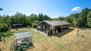 Et moderne hus med en stor patio, et glashus og en skov i baggrunden.