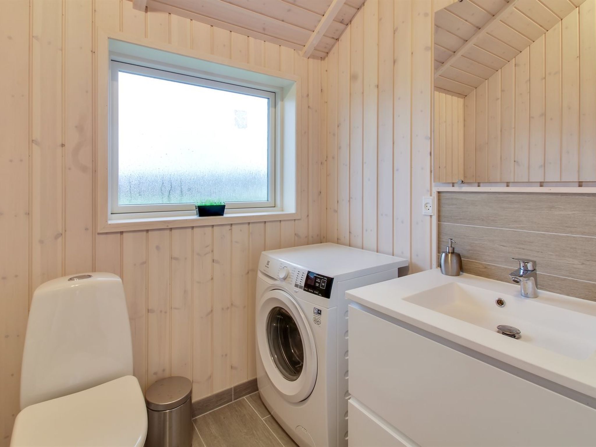 A compact, modern bathroom with wooden paneling, a washing machine, a sink, and a toilet.