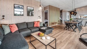 Un salon moderne avec un canapé d’angle, un coin repas avec une table et des chaises en bois, et une cuisine en arrière-plan.