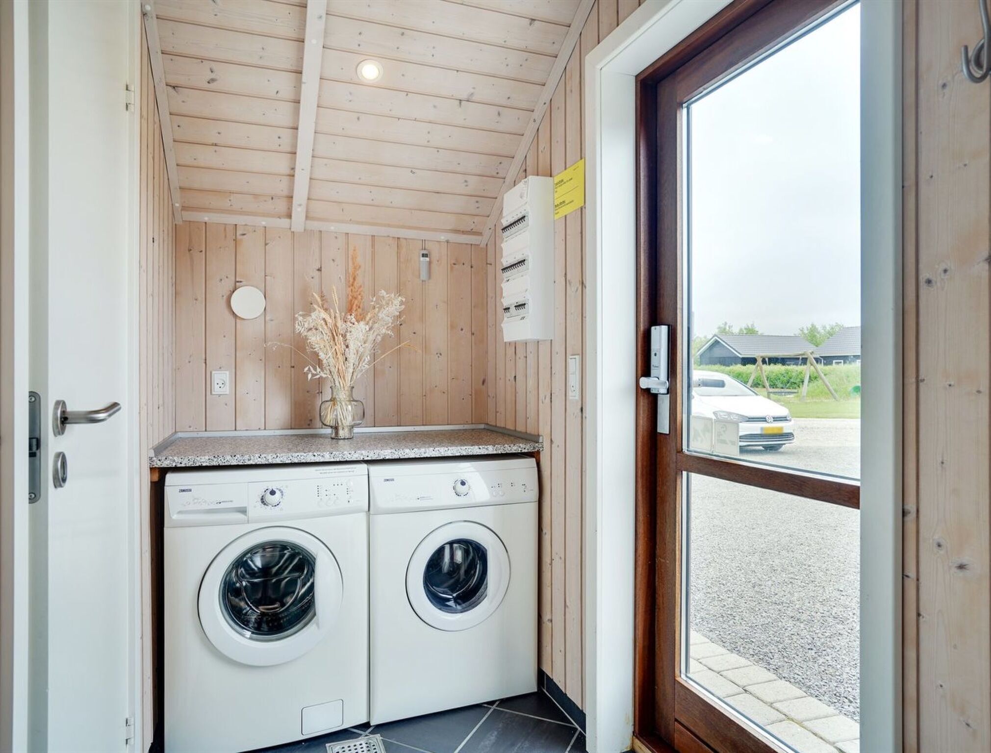 A laundry room with two front-loading washing machines, a wooden ceiling, a window with a view of a parking area, and a door with a handle.