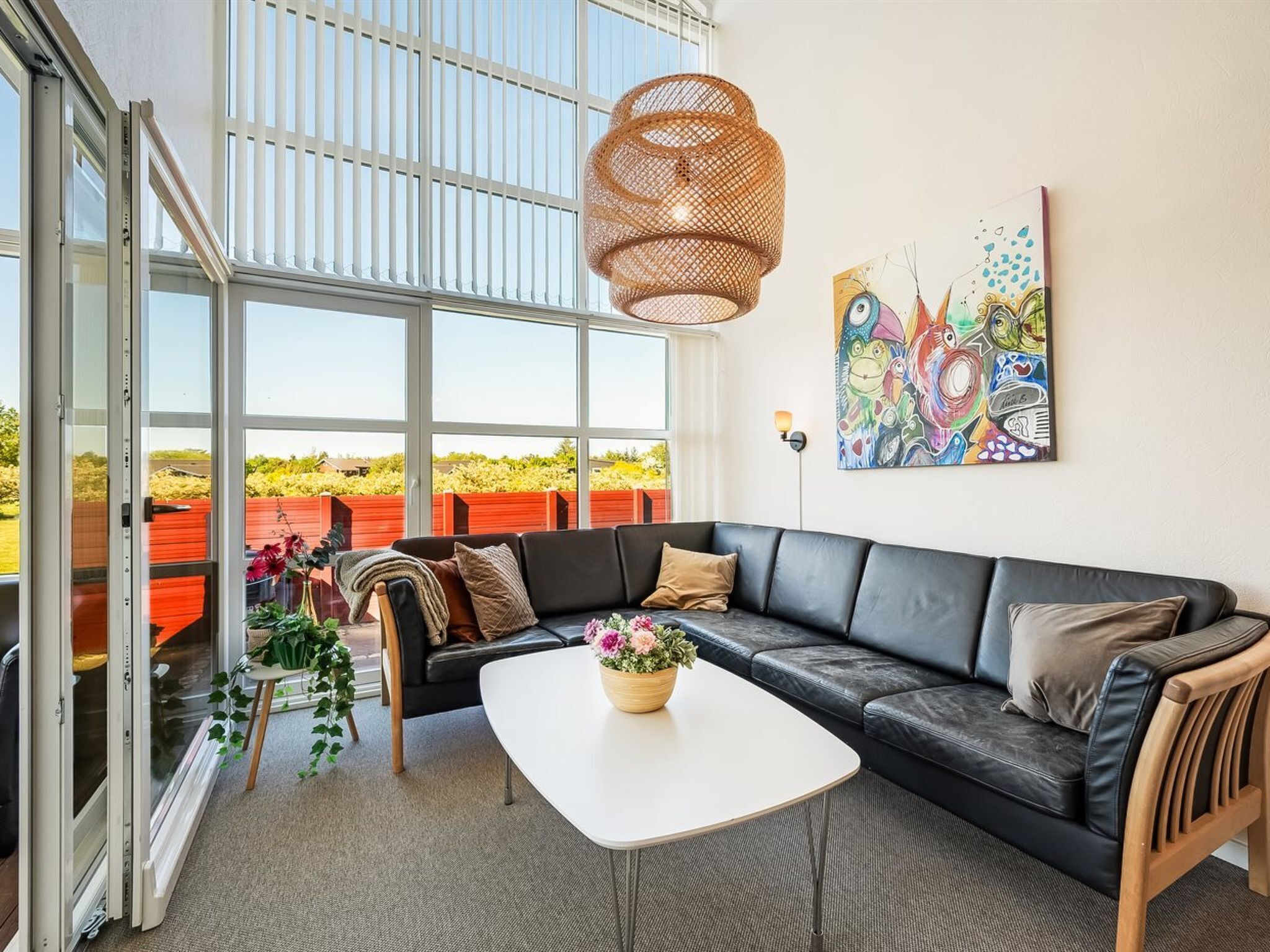 A modern living room with a black leather sofa, a white coffee table, and a large window with a view of a red fence and greenery.
