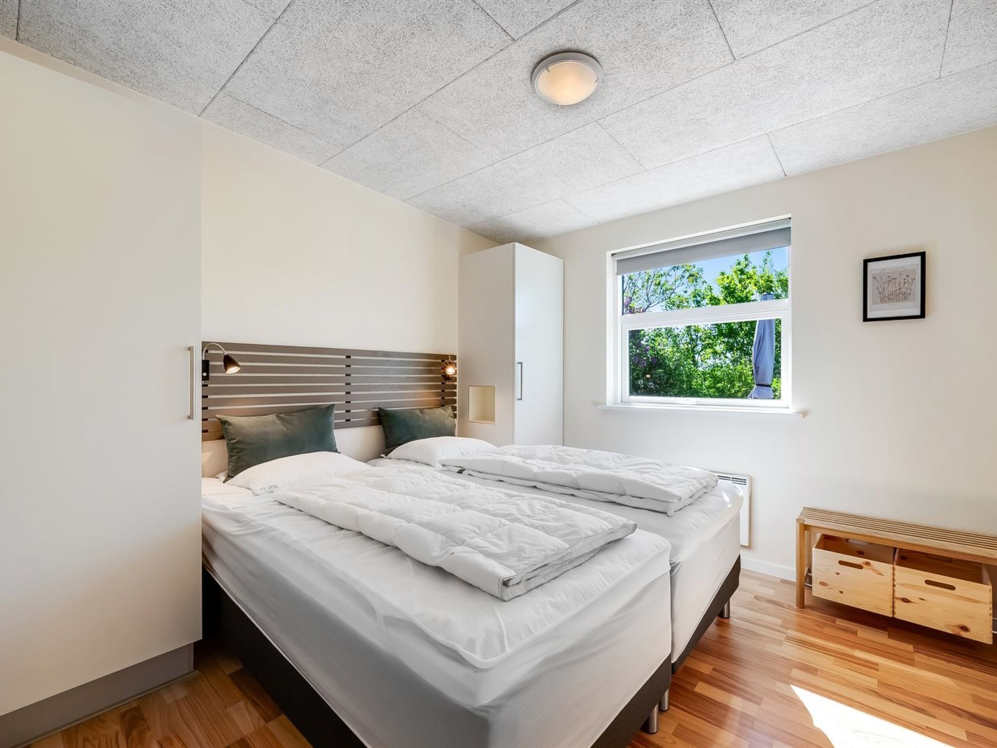 Une chambre moderne avec deux lits, une tête de lit en bois, une table de chevet avec une lampe, une fenêtre donnant sur un paysage verdoyant et un banc en bois avec des tiroirs.